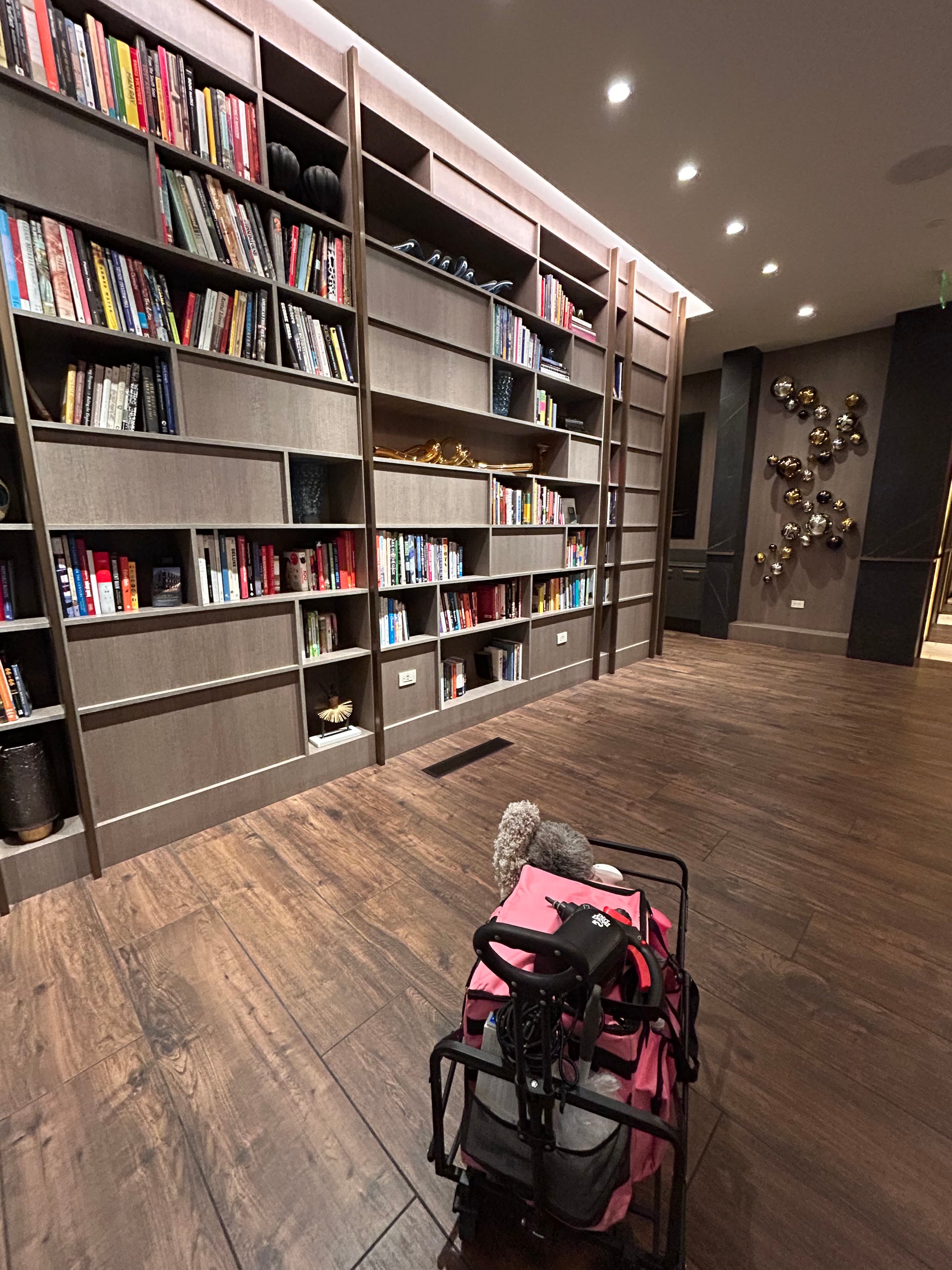 Large wooden bookshelf filled with books and a pink cleaning cart on dark wood floors.