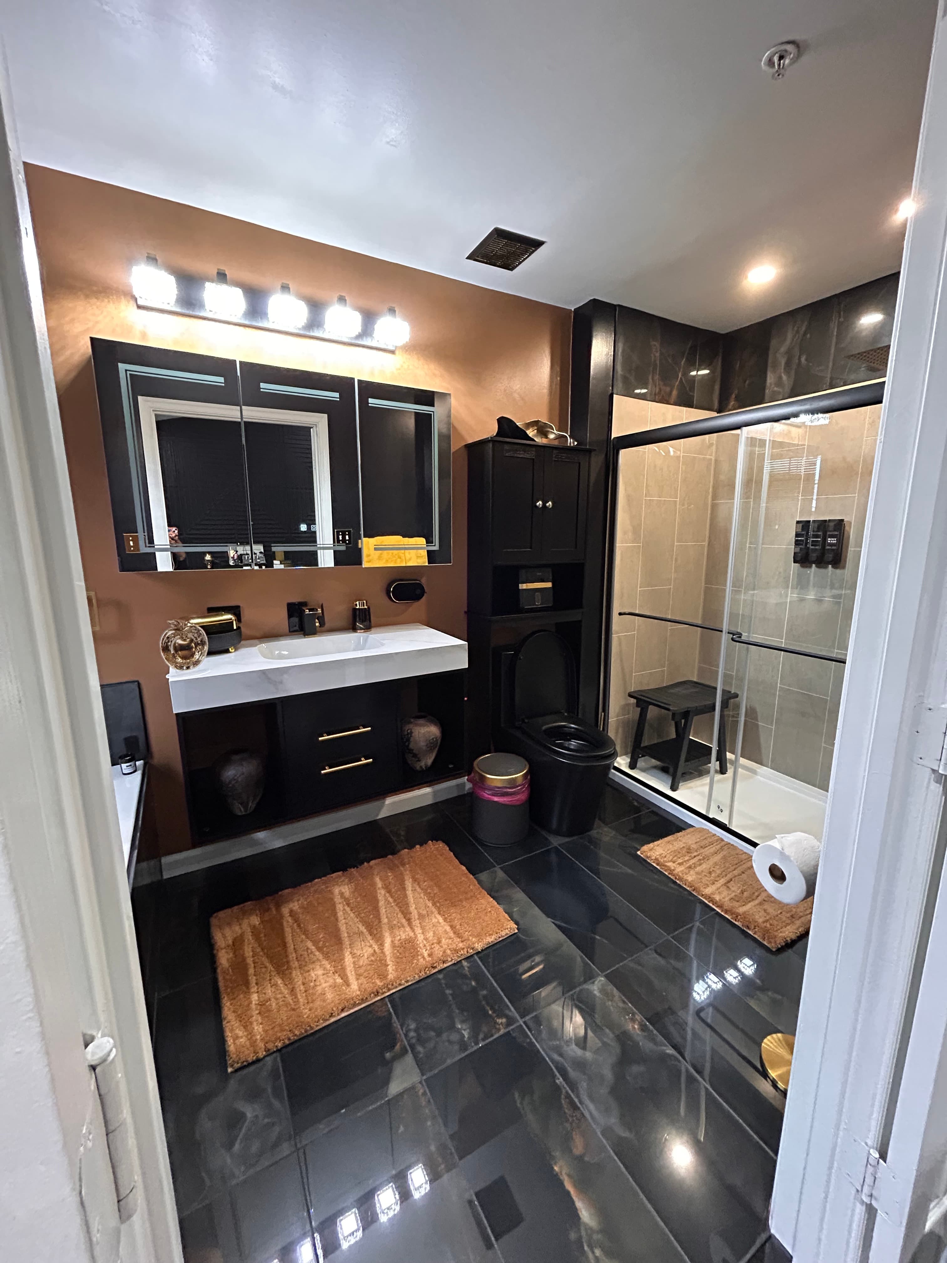 Modern bathroom featuring copper walls, black vanity, black toilet, and glass-enclosed walk-in shower.
