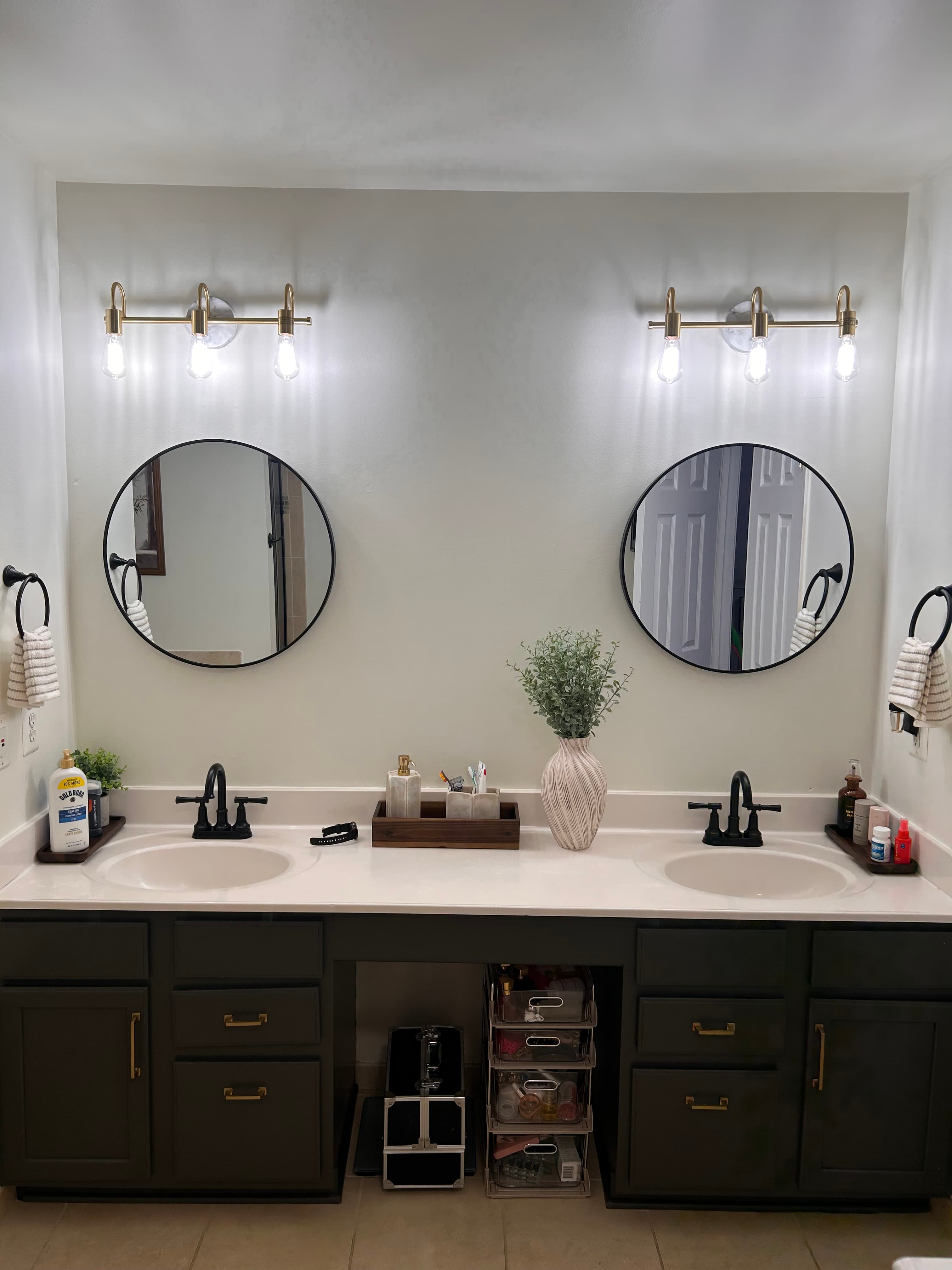 Modern double vanity with dark green cabinets, gold hardware, round mirrors, and gold lighting.